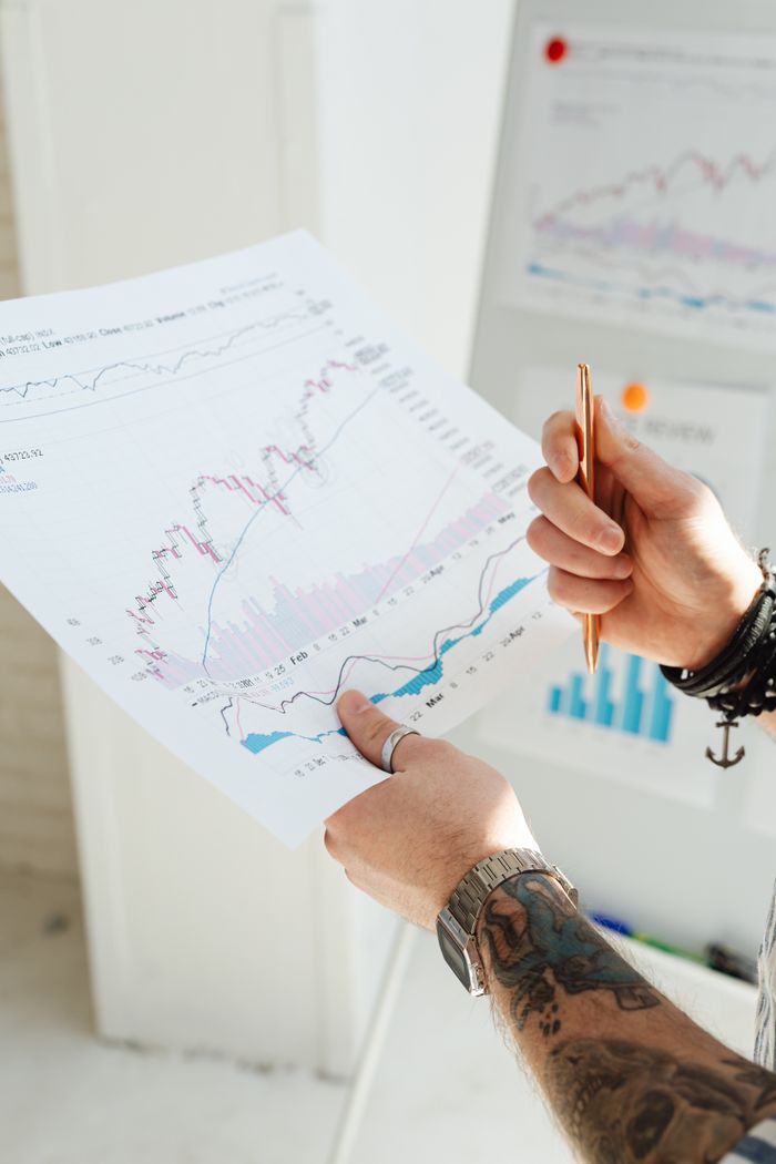 gallery-6 Close-up of tattooed hands holding financial charts and pen, illustrating market analysis.