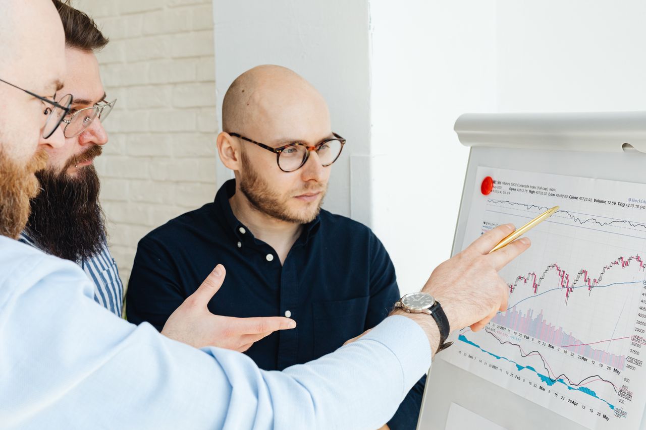 about-01 Three men discussing financial charts on a whiteboard during a business meeting.